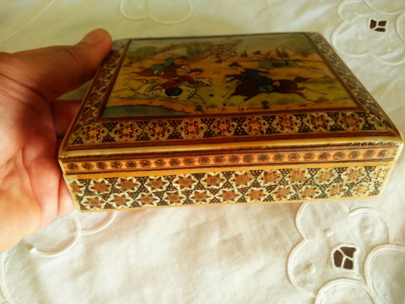 May include: A rectangular wooden box with a detailed painting on the lid depicting figures on horseback in a landscape. The box has intricate geometric patterns in shades of brown, gold, and cream. The box is held by a hand.