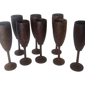 Op de afbeelding: Een set van zeven hoge, slanke bekers gemaakt van donkerbruin hout met een zichtbare, getextureerde nerf. Elke beker heeft een diepe, ronde kom, een dunne steel en een ronde voet, rechtopstaand in een gegroepeerde opstelling.