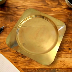 Op de afbeelding: Een goudkleurige, vierkante decoratieve plaat met afgeronde hoeken. De plaat heeft een cirkelvormige inkeping in het midden. De plaat is gemaakt van metaal en heeft een glanzende afwerking. De plaat ligt op een houten oppervlak.