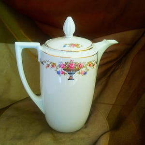 May include: White porcelain coffee pot with a floral design. The pot has a lid with a knob in the shape of a flower. The floral design features pink, blue, and yellow flowers.