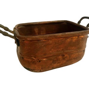 Puede incluir: Una olla rectangular de cobre con dos asas arqueadas. La olla tiene una base redondeada y una superficie ligeramente texturizada. El cobre tiene un color marrón rojizo cálido, lo que sugiere que es un artículo antiguo o vintage. Ideal para la decoración de la cocina.