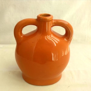 Può includere: Una brocca in ceramica color arancione brillante e lucida con un corpo arrotondato, un collo stretto e due manici arcuati simmetrici fissati ai lati del collo. La superficie presenta una finitura liscia e riflettente di colore uniforme.