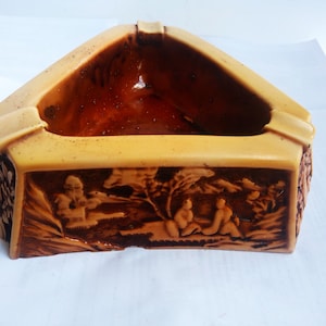 May include: A triangular ceramic ashtray with a brown interior and a beige exterior. The exterior is decorated with a relief design of people and trees.