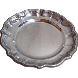Peut inclure: Une assiette argentée avec un bord festonné. L'assiette a une surface lisse et réfléchissante et un centre légèrement surélevé. L'assiette décorative est ronde et semble être en métal.