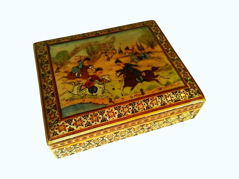 May include: A square, decorative box with a detailed painting on the lid depicting a polo match. The box has a gold-colored border with intricate floral and geometric patterns. The painting features figures on horseback in a landscape setting.