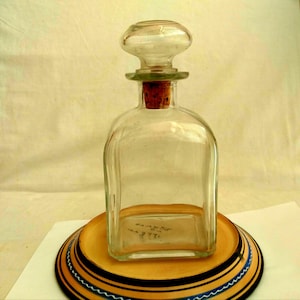 Puede incluir: Una jarra de vidrio transparente con tapón redondeado y corcho. La botella cuadrada se encuentra sobre un plato decorativo con rayas negras, azules y amarillas. La jarra está vacía.