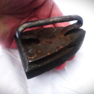 Puede incluir: Una plancha vintage de hierro fundido, con un mango negro y una base oxidada de color gris.