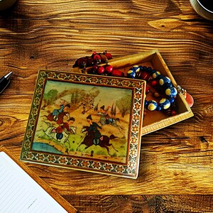 May include: An open, rectangular wooden box with a decorative lid featuring a detailed painting of horsemen in a landscape. The box contains colorful beads and other small items. The scene is set on a wooden surface.