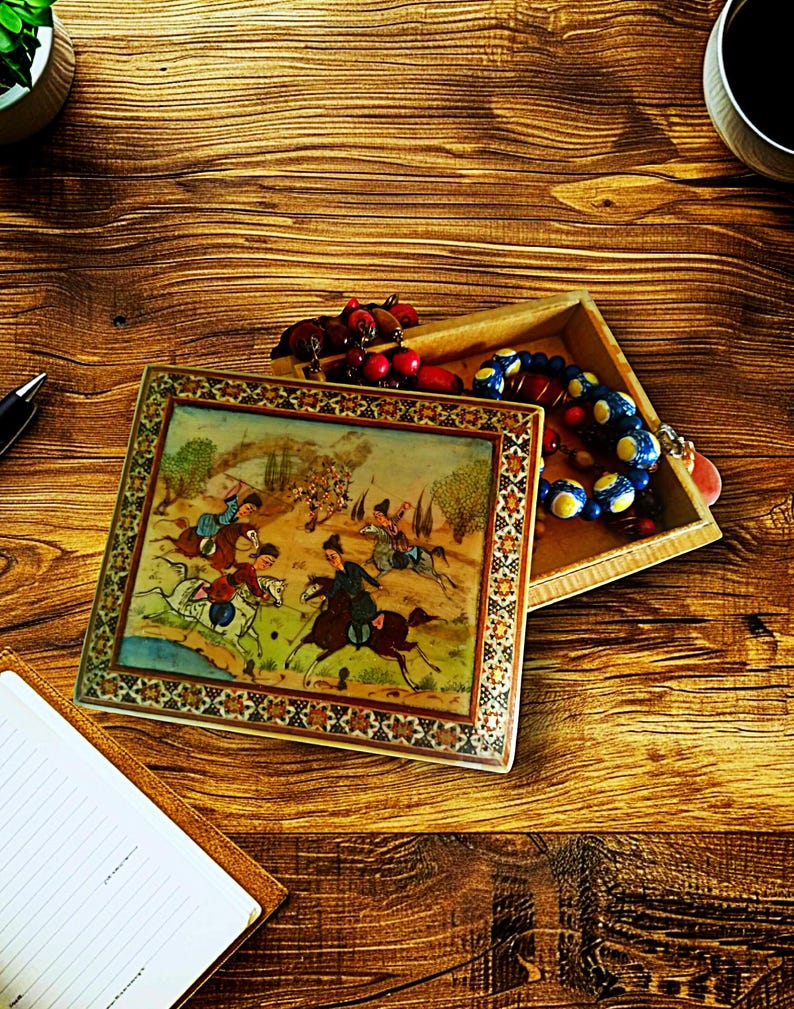 May include: An open, rectangular wooden box with a decorative lid featuring a detailed painting of horsemen in a landscape. The box contains colorful beads and other small items. The scene is set on a wooden surface.