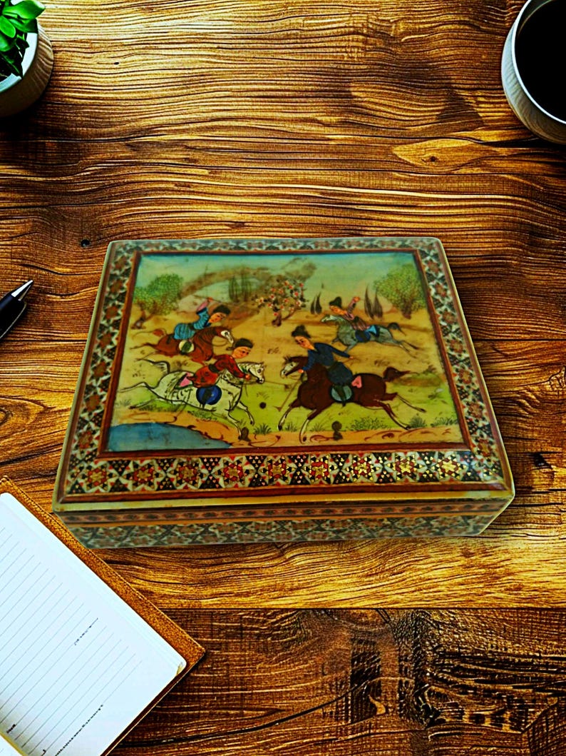 May include: A rectangular wooden box with a detailed painting on the lid depicting a polo match with riders on horseback. The box has a decorative border with intricate patterns and is set on a wooden surface.