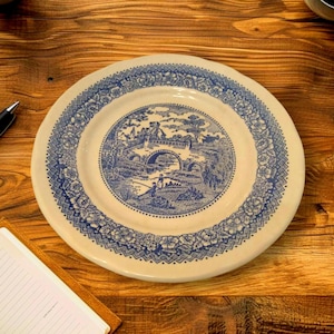 Può includere: Un piatto rotondo in ceramica bianco sporco con un disegno blu. L'immagine centrale raffigura un ponte su un fiume con figure, circondato da un bordo floreale. Il piatto è appoggiato su una superficie di legno.