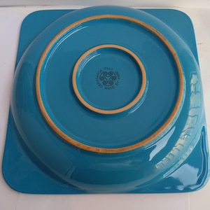 Op de afbeelding: Een turquoise keramische plaat met een vierkante basis en een cirkelvormig midden. De plaat heeft een opstaande rand met een bruine rand. De onderkant van de plaat heeft de tekst "CASASARDA ITALY HAND MADE BY MIN".