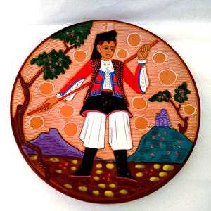 Op de afbeelding: Een ronde, decoratieve plaat met een volkskunstontwerp. De plaat toont een figuur in traditionele klederdracht, met een staf, tegen een landschap met bomen en bergen. Het kleurenpalet omvat rood, zwart, wit en oranje.