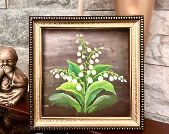May include: A framed painting of lily of the valley flowers. The artwork features white bell-shaped flowers with green leaves against a brown background. The frame is gold with a beaded edge. The painting measures 13x13 cm.