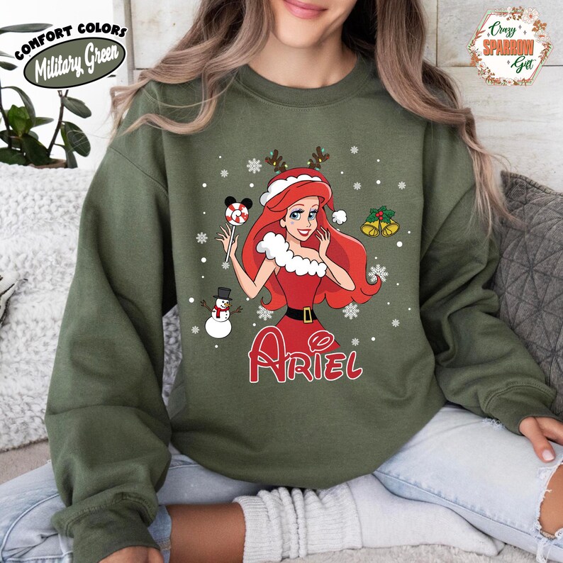 May include: Military green sweatshirt featuring a Christmas-themed Ariel graphic. The design includes snowflakes, a snowman, and a candy cane. The word "Ariel" is written in red script. The sweatshirt has the words "Comfort Colors" and "Military Green" printed on it.