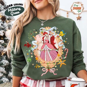 May include: Military green sweatshirt featuring a cartoon illustration of Ariel from The Little Mermaid in a Christmas-themed design. The design includes a decorative frame, lights, and sea creatures. The text "Gildan Colors Military Green" is visible.