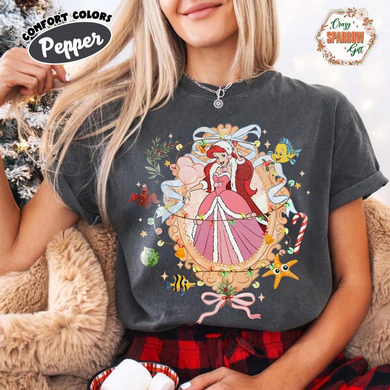 Puede incluir: Camiseta gris oscuro con una ilustraci&oacute;n festiva de una princesa con un vestido rosa y un gorro de Pap&aacute; Noel, rodeada de decoraciones navide&ntilde;as. La camiseta tiene el texto "Comfort Colors Pepper" en la parte superior izquierda.