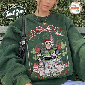May include: Forest green sweatshirt featuring a festive design with Buzz Lightyear in a Santa hat, surrounded by Christmas decorations and a castle. The sweatshirt has the text "Gildan Colors Forest Green" on the upper left.