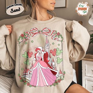May include: Sand-coloured sweatshirt with a festive illustration of a princess in a Christmas design. The graphic includes a castle, Mickey Mouse balloons, and a decorative bow. The sweatshirt has the text "Gildan Colors Sand".