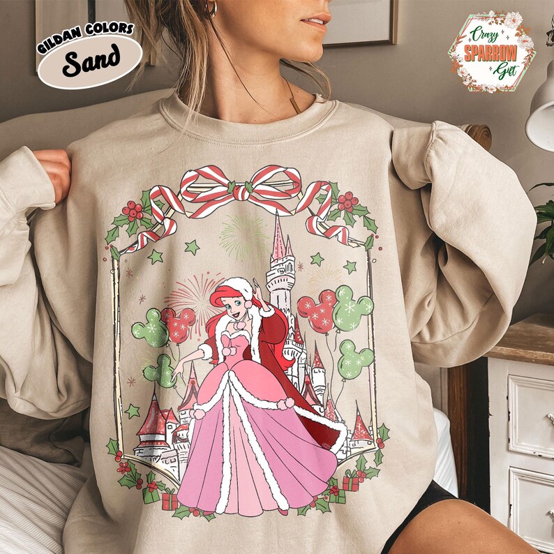May include: Sand-coloured sweatshirt with a festive illustration of a princess in a Christmas design. The graphic includes a castle, Mickey Mouse balloons, and a decorative bow. The sweatshirt has the text "Gildan Colors Sand".