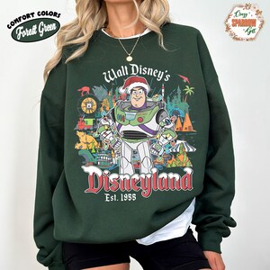K&ouml;nnte beinhalten: Forstgr&uuml;nes Sweatshirt mit einer Grafik von Buzz Lightyear mit Weihnachtsm&uuml;tze vor Disneyland-Bildern. Der Text "Walt Disney's Disneyland Est. 1955" ist in Rot und Wei&szlig; gehalten. Das Sweatshirt hat die Worte "Comfort Colors" und "Forest Green" in einem wei&szlig;en Oval.