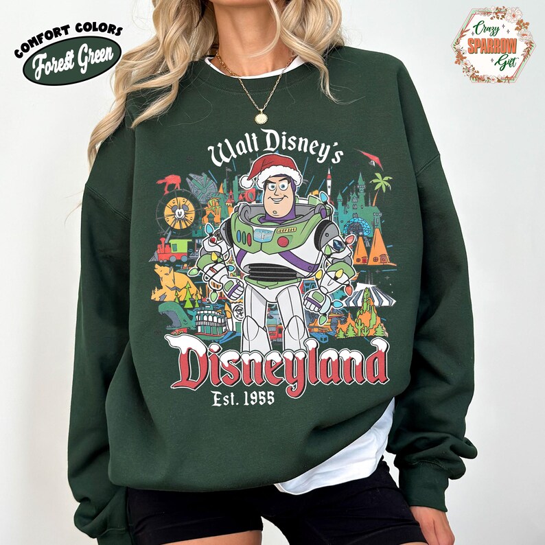 K&ouml;nnte beinhalten: Forstgr&uuml;nes Sweatshirt mit einer Grafik von Buzz Lightyear mit Weihnachtsm&uuml;tze vor Disneyland-Bildern. Der Text "Walt Disney's Disneyland Est. 1955" ist in Rot und Wei&szlig; gehalten. Das Sweatshirt hat die Worte "Comfort Colors" und "Forest Green" in einem wei&szlig;en Oval.