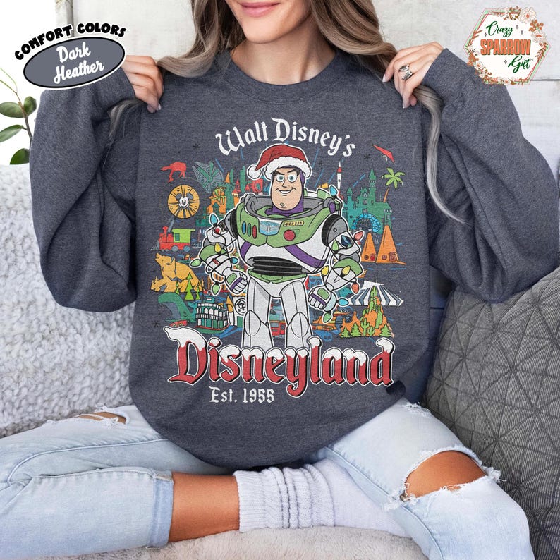K&ouml;nnte beinhalten: Dunkelgraues Sweatshirt mit einem Buzz Lightyear-Motiv mit Weihnachtsm&uuml;tze, vor dem Hintergrund von Disneyland. Der Text lautet "Walt Disney's Disneyland Est. 1955". Das Sweatshirt hat einen Rundhalsausschnitt und lange &Auml;rmel.
