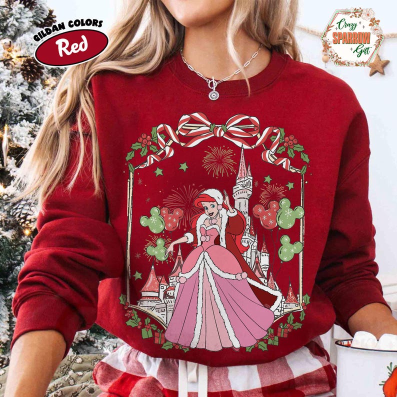 May include: Red sweatshirt featuring a princess in a pink gown, a castle, and fireworks. The design includes a bow and holly at the top. The text "Gildan Colors Red" is in a red oval on the top left.