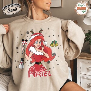 May include: Sand-colored Comfort Colors sweatshirt featuring a Christmas-themed Ariel design. The graphic includes Ariel in a Santa hat and red dress, holding a candy cane. Snowflakes, a snowman, and bells complete the festive design. The word "Ariel" is written in red.