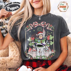 May include: Dark gray t-shirt featuring a Christmas-themed Buzz Lightyear graphic. The design includes a castle, fireworks, and a festive wreath. The shirt has the words "Comfort Colors Pepper" on the top left.