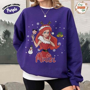 May include: A purple sweatshirt featuring a graphic of Ariel from The Little Mermaid in a Christmas-themed design. The design includes snowflakes, a snowman, and the name "Ariel" in red. The sweatshirt has the words "Comfort Colors" and "Purple" on the upper left.