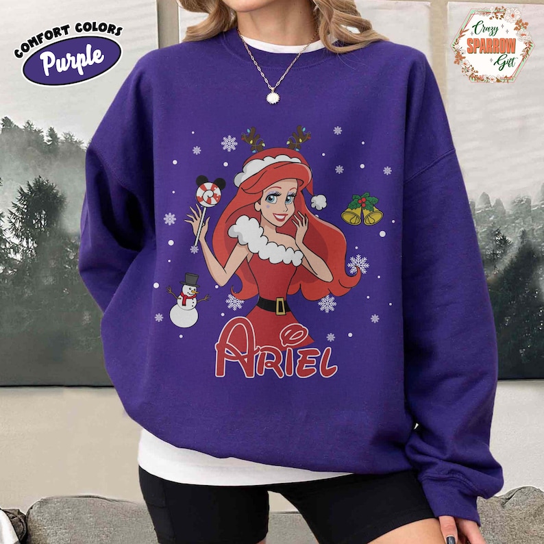 May include: A purple sweatshirt featuring a graphic of Ariel from The Little Mermaid in a Christmas-themed design. The design includes snowflakes, a snowman, and the name "Ariel" in red. The sweatshirt has the words "Comfort Colors" and "Purple" on the upper left.