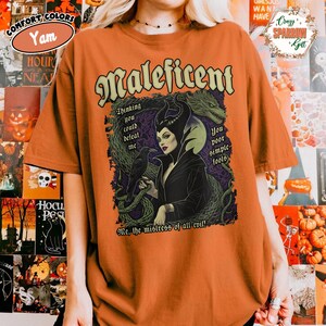 Pode incluir: Camiseta laranja com um desenho de Malévola, com texto que diz "Malévola", "Pensando que você poderia me derrotar" e "Pobres tolos". O design inclui um corvo e elementos de dragão.