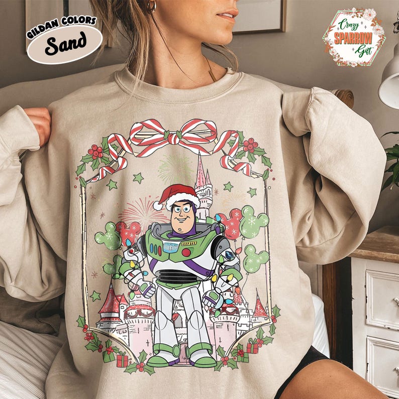 May include: Sand-colored sweatshirt featuring a festive Buzz Lightyear design. The graphic includes Buzz wearing a Santa hat, surrounded by Christmas decorations, a castle, and Mickey Mouse balloons. The sweatshirt has the text "GILDAN COLORS Sand" in the upper left corner.