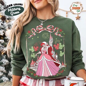 May include: Military green sweatshirt with a festive design. It features a princess in a pink dress, a castle, and Mickey Mouse silhouettes. The design includes a red and white bow, fireworks, and holly. The sweatshirt has the text "Gildan Colors Military Green".
