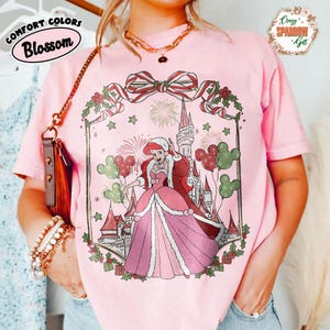 May include: Pink t-shirt featuring a princess in festive attire, holding Mickey Mouse balloons, with a castle backdrop. The shirt displays the text "Comfort Colors Blossom" and "Crazy Sparrow Gift".