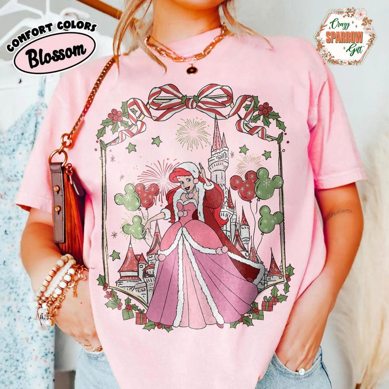 May include: Pink t-shirt featuring a princess in festive attire, holding Mickey Mouse balloons, with a castle backdrop. The shirt displays the text "Comfort Colors Blossom" and "Crazy Sparrow Gift".