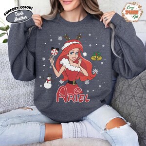 May include: Dark heather gray sweatshirt featuring a Christmas-themed Ariel graphic. The design includes Ariel in a red dress, holding a candy cane, with snowflakes and Christmas bells. The word "Ariel" is written in red.