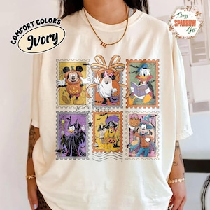 Op de afbeelding: Ivoor kleurig T-shirt met een Halloween ontwerp. Het ontwerp toont Disney-personages in kostuums, gepresenteerd als postzegels. Het T-shirt heeft de woorden "Comfort Colors" en "Ivory".