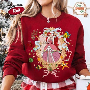 Puede incluir: Sudadera roja con una imagen enmarcada de una princesa de dibujos animados con un vestido rosa, rodeada de decoraciones festivas. La sudadera tiene el texto "Gildan Colors Red" en un &oacute;valo rojo. El dise&ntilde;o incluye una estrella de mar, un pez y un bast&oacute;n de caramelo.