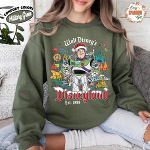 K&ouml;nnte beinhalten: Milit&auml;rgr&uuml;nes Sweatshirt mit einer Grafik von Buzz Lightyear mit Weihnachtsm&uuml;tze und Disneyland-Motiven. Der Text lautet "Walt Disney's Disneyland Est. 1955" in Rot und Wei&szlig;. Auf dem Sweatshirt stehen auch die W&ouml;rter "Comfort Colors" und "Military Green".