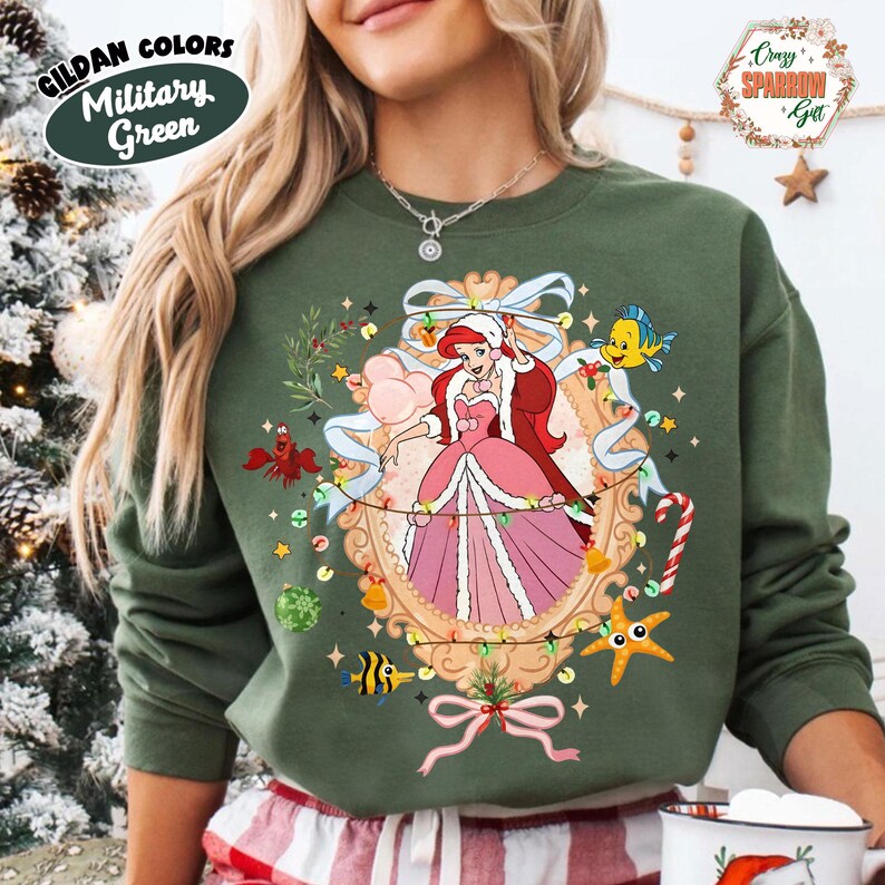Puede incluir: Sudadera verde militar con un dise&ntilde;o navide&ntilde;o que presenta a Ariel de La Sirenita. La ilustraci&oacute;n incluye un marco decorativo, luces y criaturas marinas. La sudadera tiene el texto "Gildan Colors Military Green".