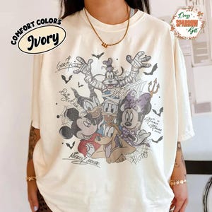 May include: Ivory-coloured t-shirt with a Halloween-themed graphic of Disney characters, including Mickey Mouse, Minnie Mouse, Goofy, Donald Duck, and Pluto. The shirt has the words "Comfort Colors" and "Ivory" printed on it.
