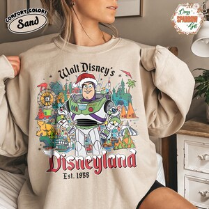 K&ouml;nnte beinhalten: Sandfarbenes Sweatshirt mit Disneyland-Design. Die Grafik zeigt Buzz Lightyear mit Weihnachtsm&uuml;tze, umgeben von Disneyland-Bildern. Der Text lautet "Walt Disney's Disneyland Est. 1955". Das Sweatshirt hat einen Rundhalsausschnitt und lange &Auml;rmel.
