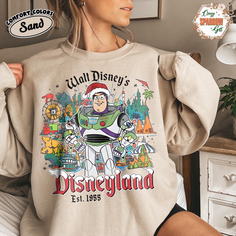K&ouml;nnte beinhalten: Sandfarbenes Sweatshirt mit Disneyland-Design. Die Grafik zeigt Buzz Lightyear mit Weihnachtsm&uuml;tze, umgeben von Disneyland-Bildern. Der Text lautet "Walt Disney's Disneyland Est. 1955". Das Sweatshirt hat einen Rundhalsausschnitt und lange &Auml;rmel.
