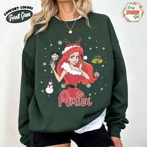 May include: Forest Green sweatshirt featuring a Christmas-themed Ariel graphic. The design includes Ariel in a red dress, holding a lollipop, with snowflakes and a snowman. The word "Ariel" is written in red script below the character.