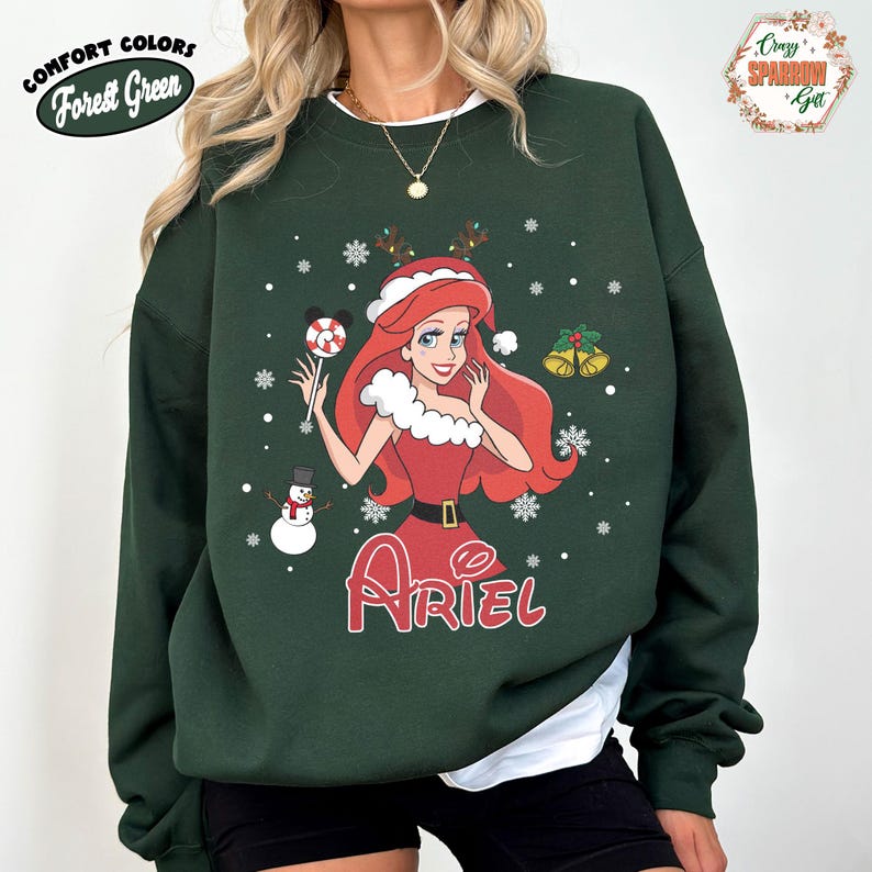 May include: Forest Green sweatshirt featuring a Christmas-themed Ariel graphic. The design includes Ariel in a red dress, holding a lollipop, with snowflakes and a snowman. The word "Ariel" is written in red script below the character.