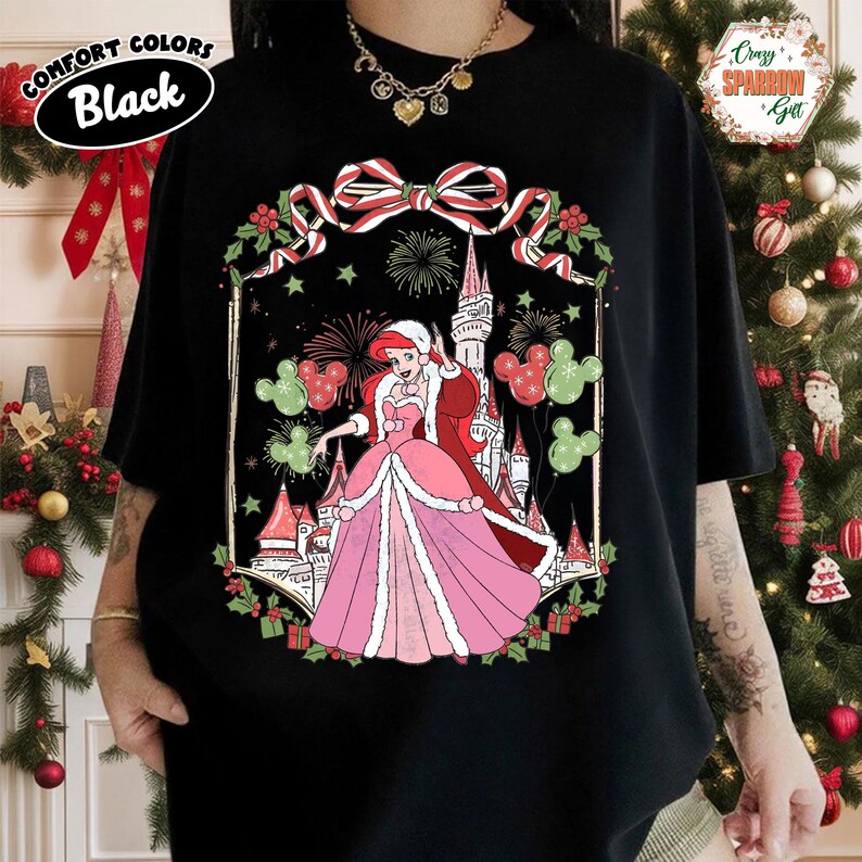 May include: Black Comfort Colors t-shirt with a festive illustration of a princess in a pink gown and red coat, with a castle backdrop, Mickey Mouse balloons, and a Christmas bow. The shirt has the text "Comfort Colors Black" in a white oval.