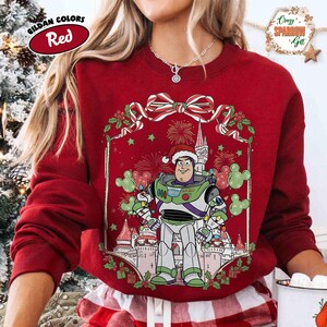 May include: Red sweatshirt featuring a graphic of Buzz Lightyear in a Santa hat, set against a castle and fireworks. The sweatshirt has a festive bow and holly design. The text "Gildan Colors Red" is in a red oval.