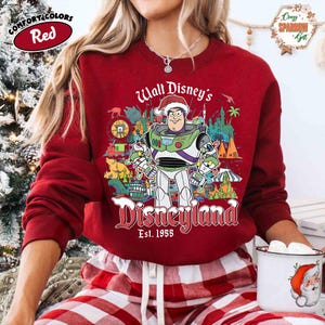 K&ouml;nnte beinhalten: Ein rotes Comfort Colors Sweatshirt mit Buzz Lightyear in einem Weihnachtsmannhut und der Aufschrift "Walt Disney's Disneyland Est. 1955". Die Grafik enth&auml;lt Disneyland-Symbole. Das Sweatshirt wird mit einer rot-wei&szlig; karierten Pyjamahose getragen.